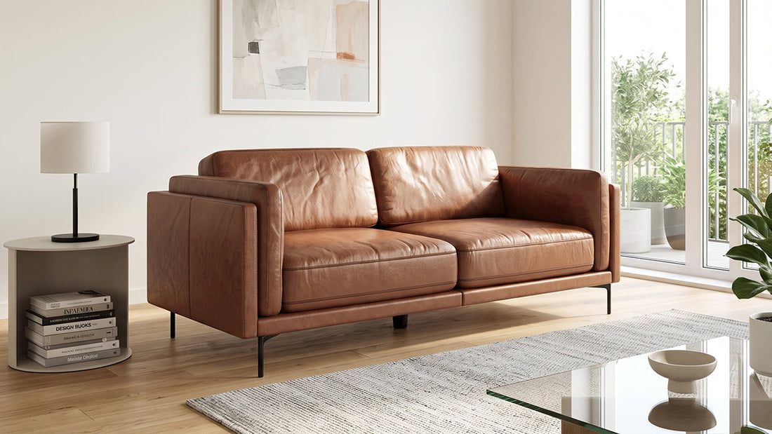 Tan leather two-seater sofa with slim black metal legs, positioned in a well-lit modern living room next to a side table with stacked books.