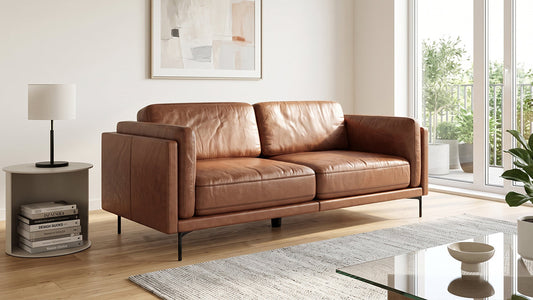 Tan leather two-seater sofa with slim black metal legs, positioned in a well-lit modern living room next to a side table with stacked books.