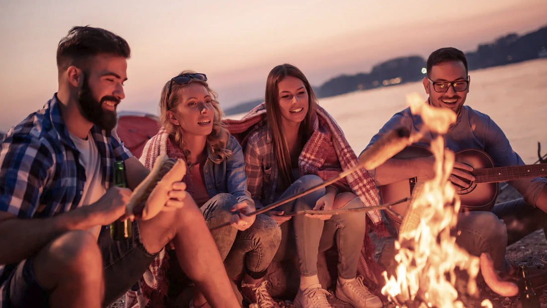 Friends gathered around a cozy fire pit on the beach at sunset, roasting food and playing guitar, illustrating what to do with your outdoor fire pit.