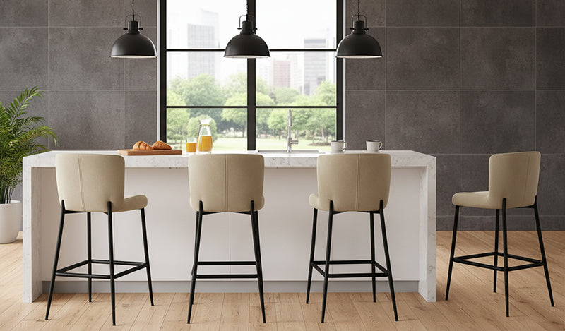 A modern kitchen island with a white countertop and base, paired with four bar stools featuring cream-colored upholstered seats and slim, matte black metal legs, set against a dark gray tiled wall with large windows in a bright room.
