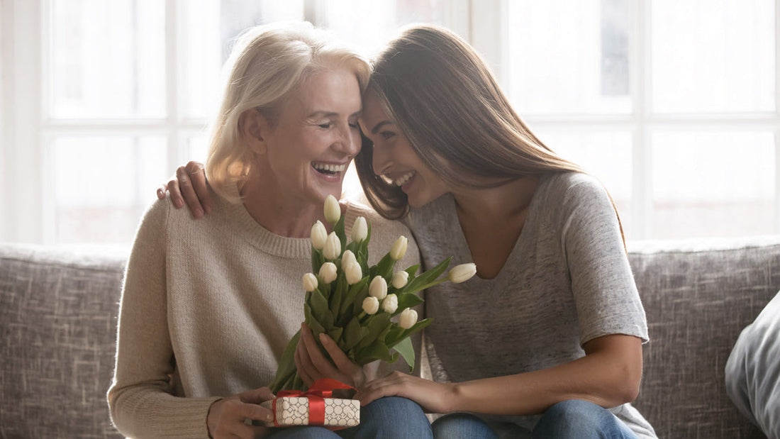 Mother and daughter hugging and smiling while holding a bouquet of tulips and a small gift box, illustrating the Mother's Day furniture gift guide.