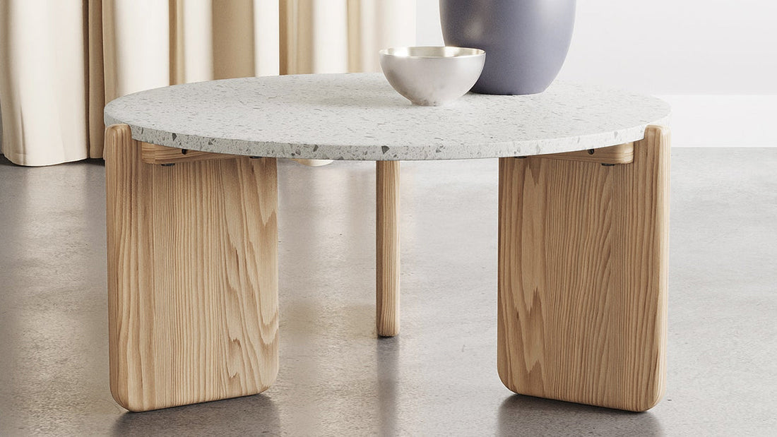 Scandinavian-inspired round coffee table featuring a speckled white tabletop and natural wood panel base, styled with a simple bowl and large vase.