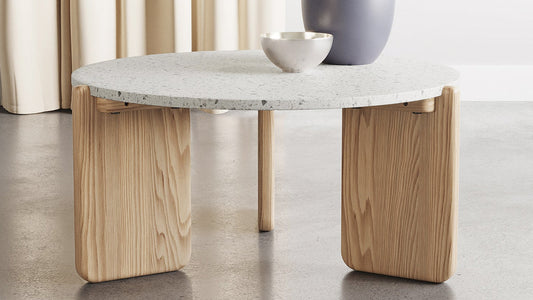 Scandinavian-inspired round coffee table featuring a speckled white tabletop and natural wood panel base, styled with a simple bowl and large vase.