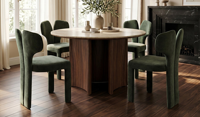 A modern dining set featuring a round, light-toned table with a dark timber, multi-panel pedestal base, surrounded by forest-green, velvet-upholstered dining chairs with a distinctive split-back design, set in a room with a dark marble fireplace.