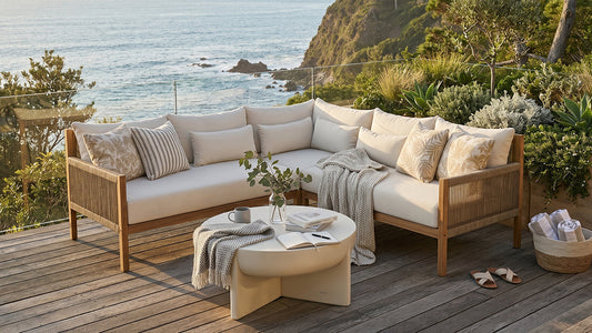 Wide-angle lifestyle shot of a beautifully styled coastal outdoor setting on a timber deck, afternoon light, with a modular lounge, teak dining table and lush planted windbreak in the background — calm, considered, and wholly inviting