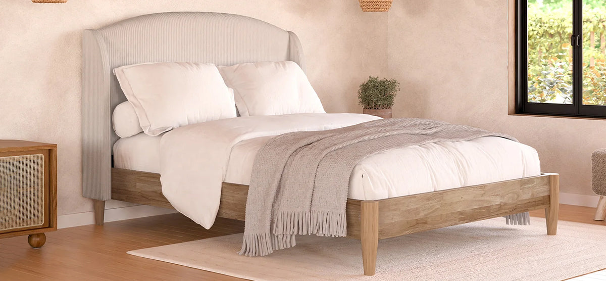 Cozy bedroom with a wooden bed frame, white linens, light-gray knit throw, upholstered headboard, woven cane nightstand, potted plant, black-framed window, and light wood flooring.