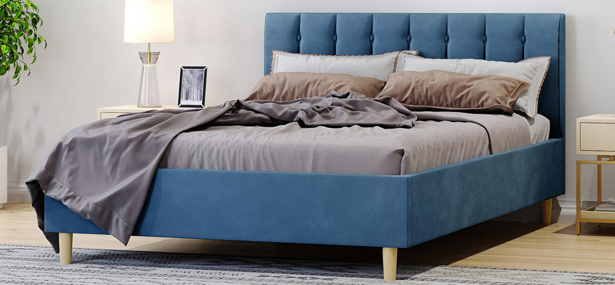 Modern bedroom with a blue upholstered platform bed featuring a tufted headboard, gray bedding, cream nightstands, gold-tone legs, a patterned rug, and a potted plant.