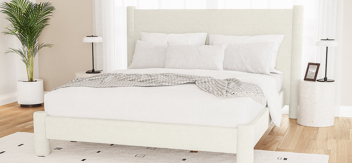 Bright bedroom with a cream-upholstered bed in white linens, mosaic-tiled nightstands with lamps, a potted palm, and sheer curtains filtering light.