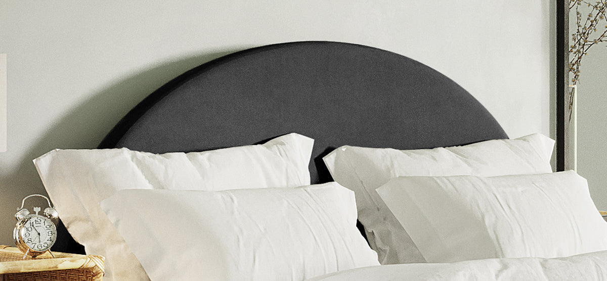 Black semi-circular upholstered headboard with four white pillows in front, adjacent to a woven bedside table featuring a small, vintage-style silver alarm clock.