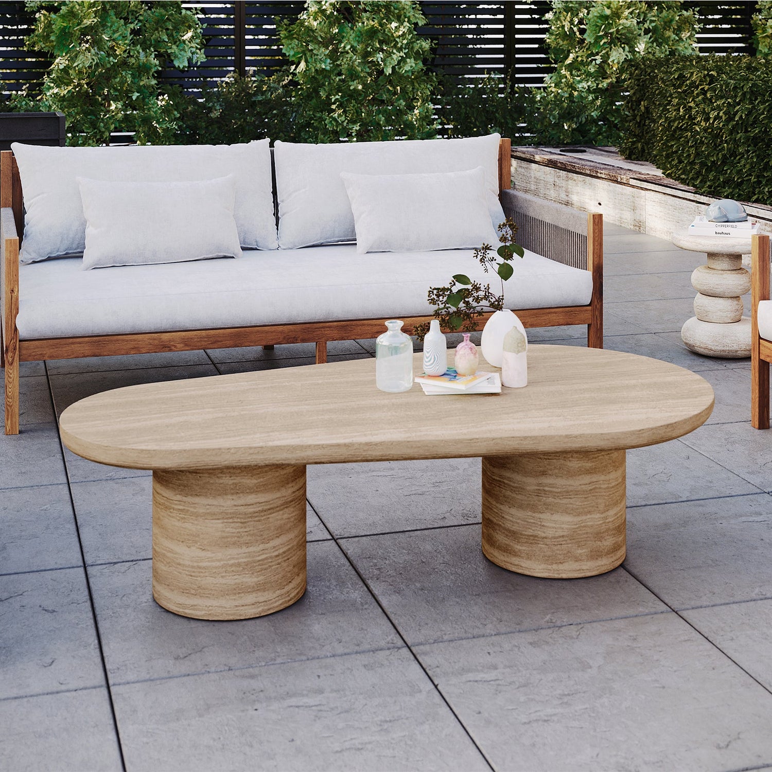 A travertine-style oval outdoor coffee table with two thick cylindrical pedestal bases, set on a grey tiled patio in front of a modern wooden sofa.