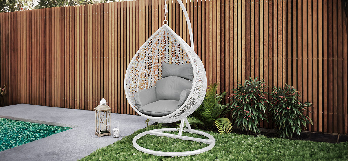 A white wicker-style hanging chair with gray cushions on a metal stand, set beside decorative lanterns, with a wooden fence and pool glimpsed in the background.
