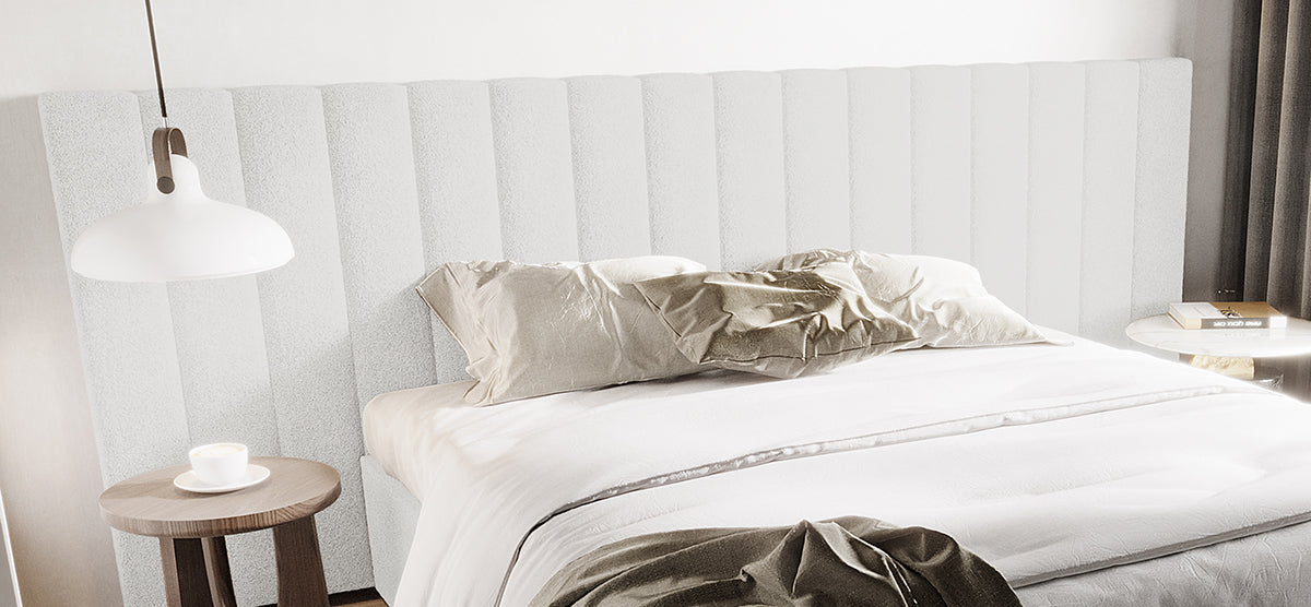Minimalist bedroom with a low upholstered platform bed featuring a channel-tufted headboard, white linens, wooden side table, pendant light, and a white table with stacked books.