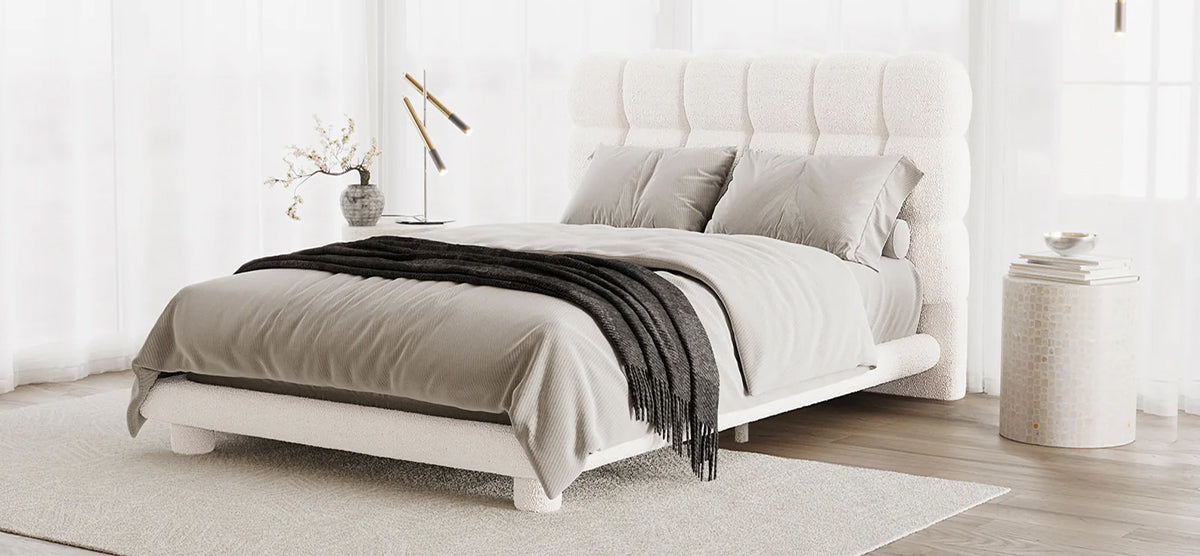 Minimalist bedroom with a bouclé-style upholstered platform bed, tall padded headboard, neutral gray bedding, sheer curtains, and a round side table with books and a decorative bowl.