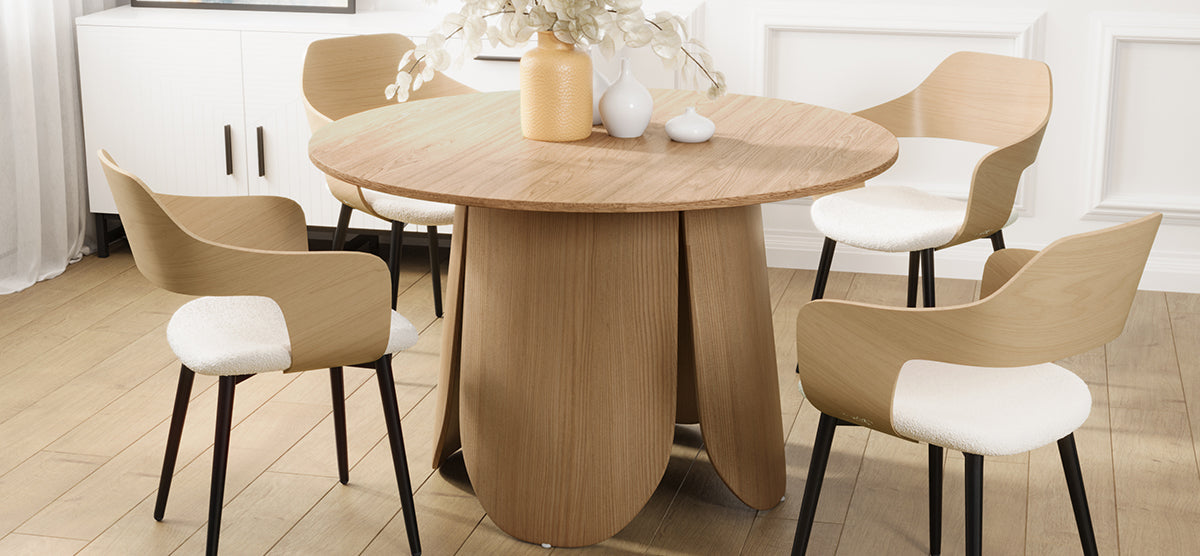 Minimalist dining nook with a round wooden table and sculptural base, four matching chairs with white cushions, and decorative vases on top. Light wood plank flooring.