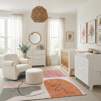 A modern nursery featuring a white crib and dresser, a cream bouclé rocking chair with matching ottoman, and an abstract-patterned rug.