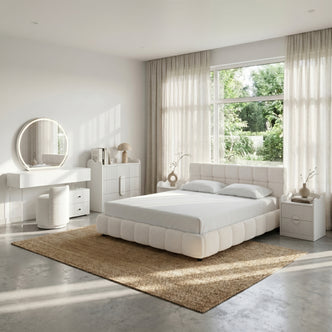 A modern bedroom with a tufted cream bouclé bed, matching bedside tables, a white vanity with a round mirror, and a large jute rug.