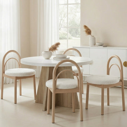 Modern round white dining table with a wooden base, surrounded by four light wood dining chairs with arched backs and white textured seats.