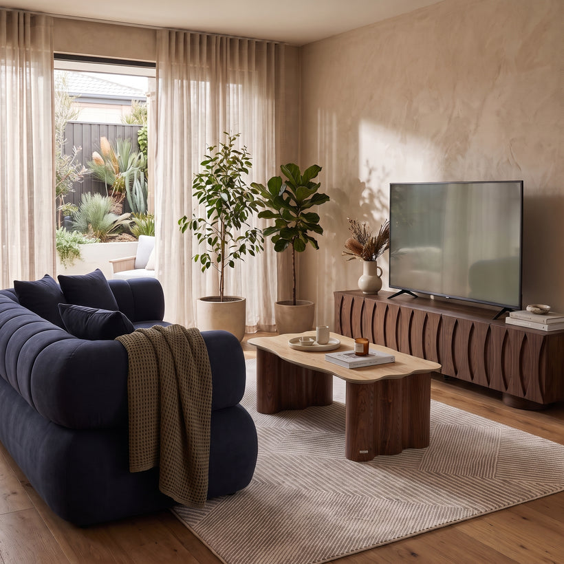 A brightly lit, modern living room featuring a plush dark blue tufted sofa with a tan throw blanket, a unique wavy-edged wooden coffee table, and a large flat-screen TV on a ribbed wood media console. A patterned beige area rug ties the space together, while two large potted plants sit by a glass door draped with sheer curtains that open to an outdoor garden patio.