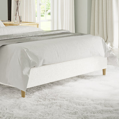 Celina Queen Boucle Fabric Bed Base in Cream White features boucle upholstery, wooden legs, and foam padding, set on a fluffy white rug with a decorative runner at the bed's foot.