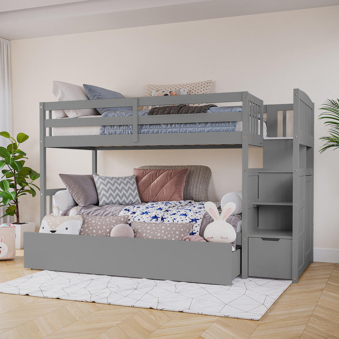 Flavia Solid Pine Wood Staircase Storage Bunk Bed in Grey features a top bunk with guardrails, a cozy trundle with cushions and toys, and a storage staircase, all on a light wood floor.
