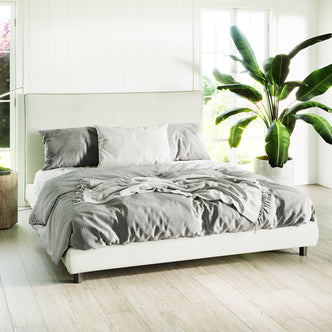 Havvan King Linen Upholstered Fabric Bed Frame - Off White in a minimalist bedroom, featuring a rumpled gray duvet, white pillow, light wood flooring, and a lush green potted plant beside it.