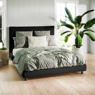 Havvan Queen Bouclé Upholstered Fabric Bed Frame - Black in a modern bedroom with a dark upholstered headboard, light gray bedding, wooden side table, desk lamp, and tropical plant by a window.