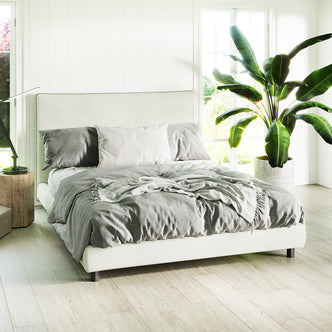Havvan Queen Linen Upholstered Fabric Bed Frame - Off White with gray bedding, two gray and one white pillow, foam-padded headboard, on light wood floor beside a simple wooden side table.