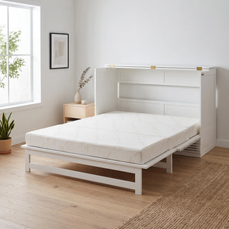 A white wooden Murphy bed folded out into a platform bed with a white patterned mattress, set in a bright room with light wood floors, a window with greenery, a small side table, and a woven rug.