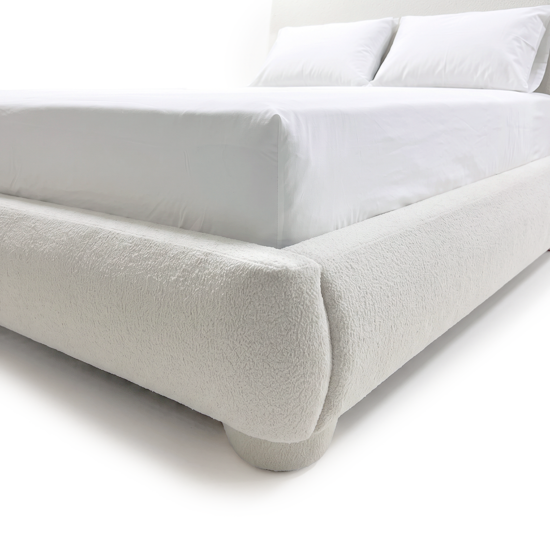 A close-up view of the white bouclé bed frame showing the rounded corner detail, upholstered side rail, and a matching rounded foot on a white background.