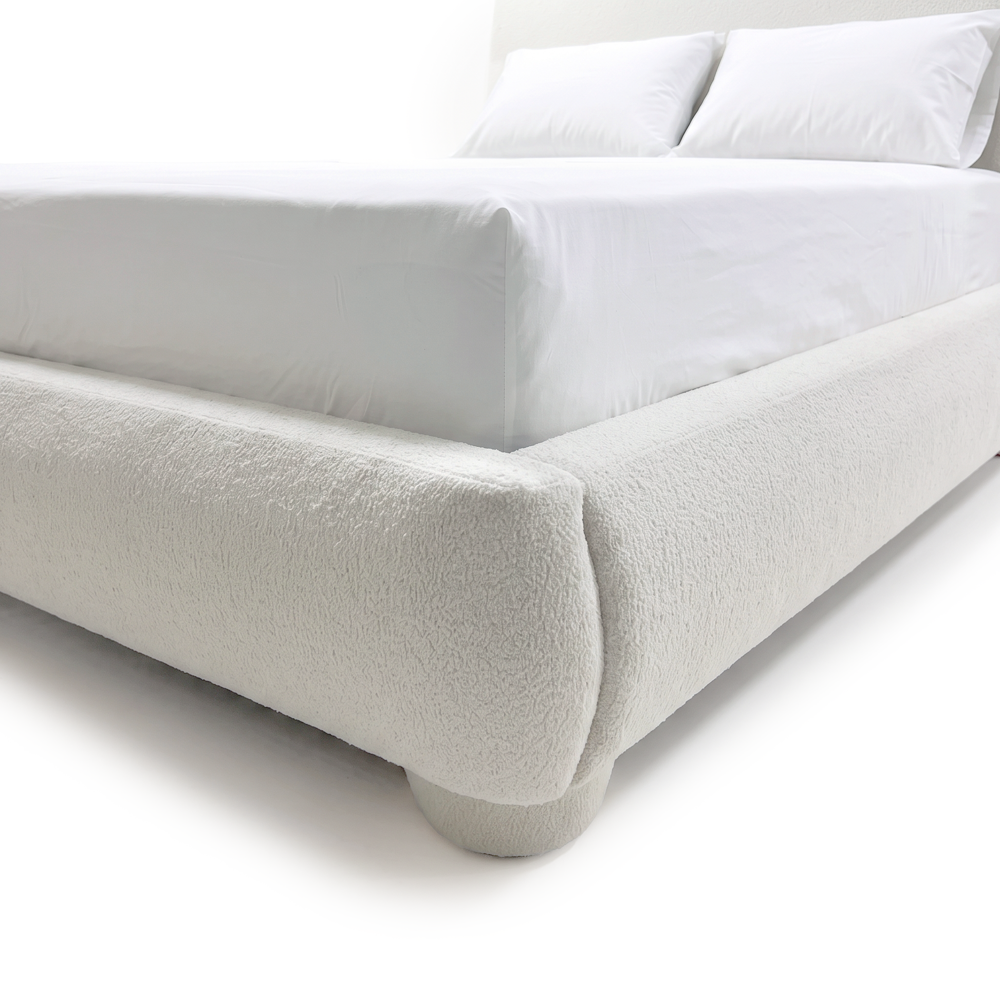 A close-up view of the white bouclé bed frame showing the rounded corner detail, upholstered side rail, and a matching rounded foot on a white background.