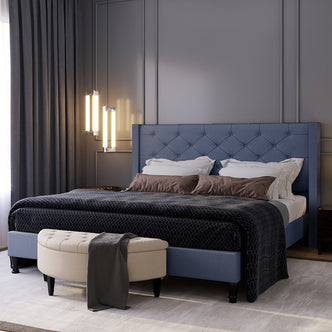 Maggie King Winged Fabric Bed Frame – Dark Blue with a tall, tufted headboard, white and brown pillows, black duvet, cream bench, and floor-length charcoal curtains in a modern bedroom setting.