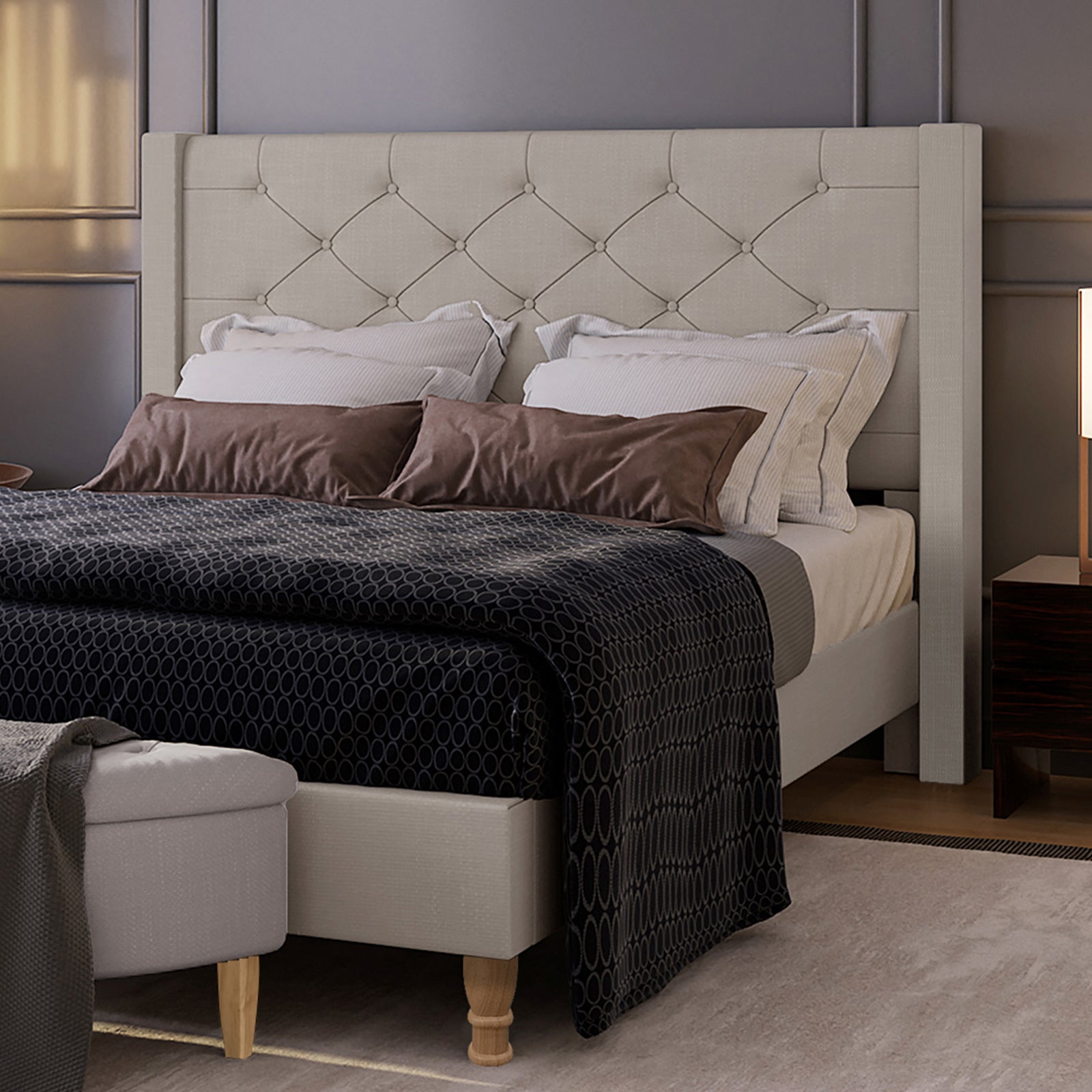 Maggie Winged Fabric Bed Frame in Warm Grey features a tall, button-tufted headboard, plush pillows, and a dark bedspread, complemented by a nightstand with a modern lamp and an upholstered bench.