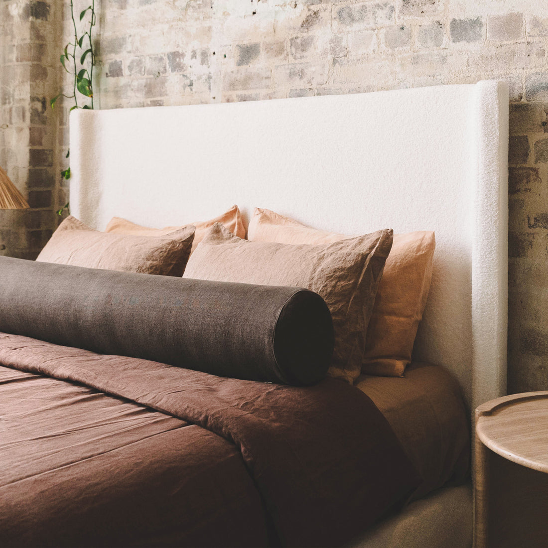 Palmira Queen Boucle Fabric Bed Frame in Cream White features a tall headboard, cream bouclé upholstery against a brick wall, complemented by a brown duvet and peachy-beige pillows.