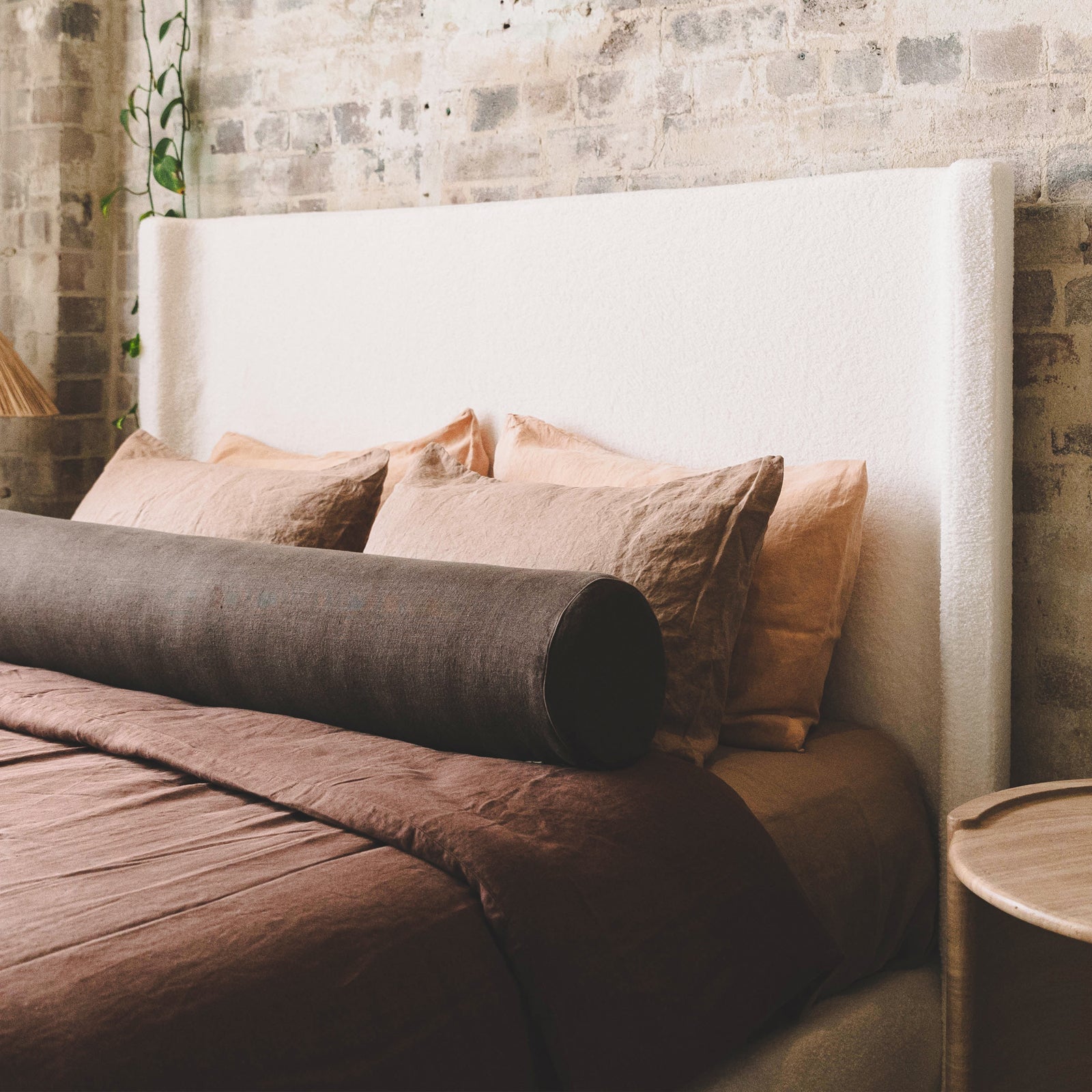 Palmira Queen Boucle Fabric Bed Frame in Cream White features a tall headboard, cream bouclé upholstery against a brick wall, complemented by a brown duvet and peachy-beige pillows.