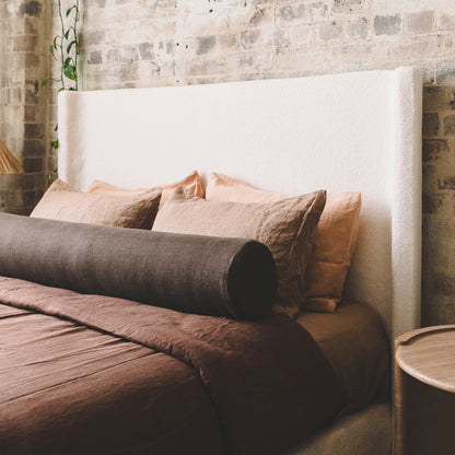 Palmira Queen Boucle Fabric Bed Frame in Cream White features a tall headboard, cream bouclé upholstery against a brick wall, complemented by a brown duvet and peachy-beige pillows.