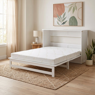 A white wooden Murphy bed folded out into a platform bed with a white patterned mattress, set in a bright room with light wood floors, a window with greenery, a small side table, and a woven rug.