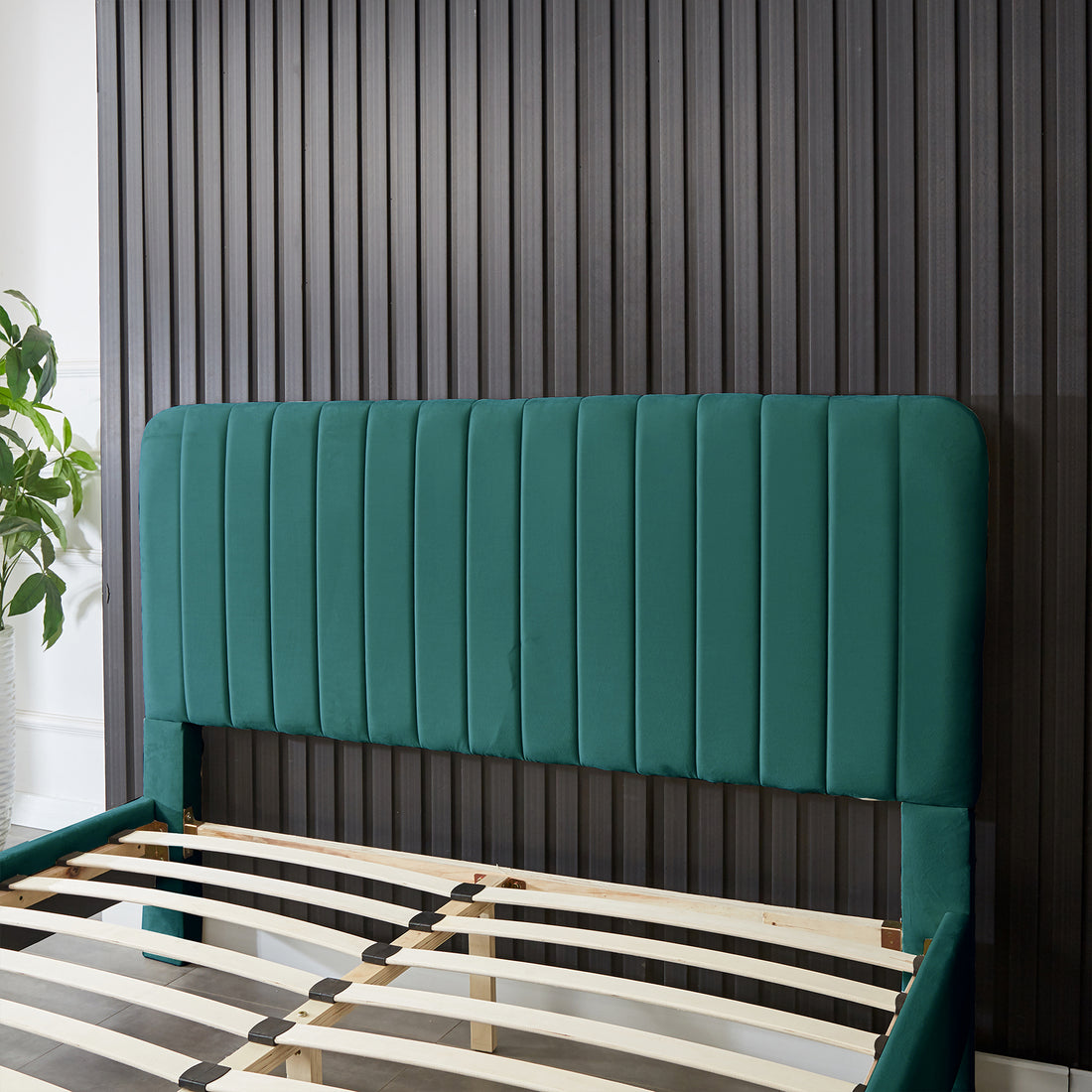 Seneca Double Upholstered Velvet Bed – Green, featuring a tall, channel-stitched headboard and deep green fabric, set against a dark wall with visible wooden slats and a leafy plant nearby.