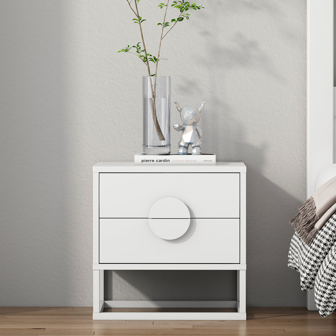 The Abigail 55cm 2-Drawer Bedside Table in white features sleek arc handles, topped with a glass vase, pierre cardin book, and silver bear figurine, complementing a minimalist bedroom setting.