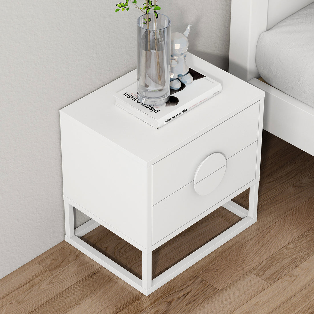 Abigail 55cm 2-Drawer Bedside Table w/ Handle - White; features sleek arc handles, two drawers, metal-frame base. Topped with pierre cardin book, glass vase, cat sculpture. Adjacent to a white bed frame.