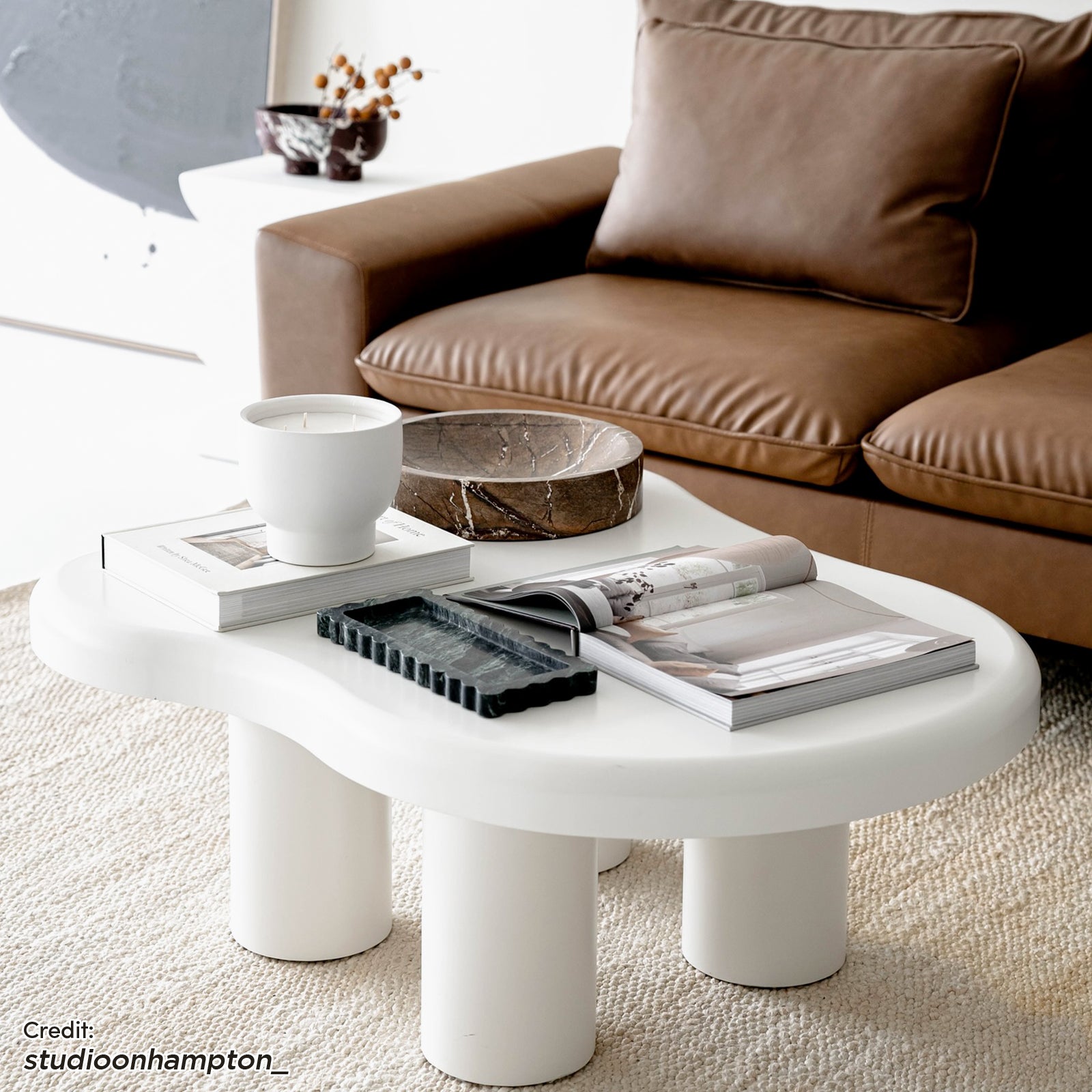 Blissey 90cm Cloud Coffee Table in White features a sculptural design with three cylindrical legs, surrounded by stylish decor, including a ceramic vessel, marble bowl, and design books on a woven rug.