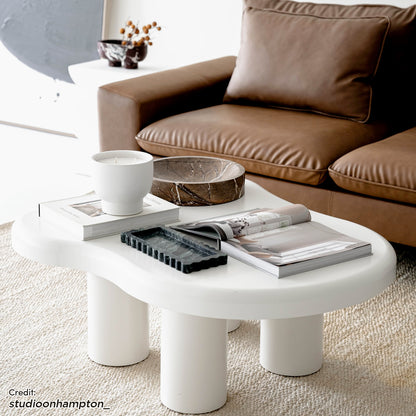 Blissey 90cm Cloud Coffee Table in White features a sculptural design with three cylindrical legs, surrounded by stylish decor, including a ceramic vessel, marble bowl, and design books on a woven rug.