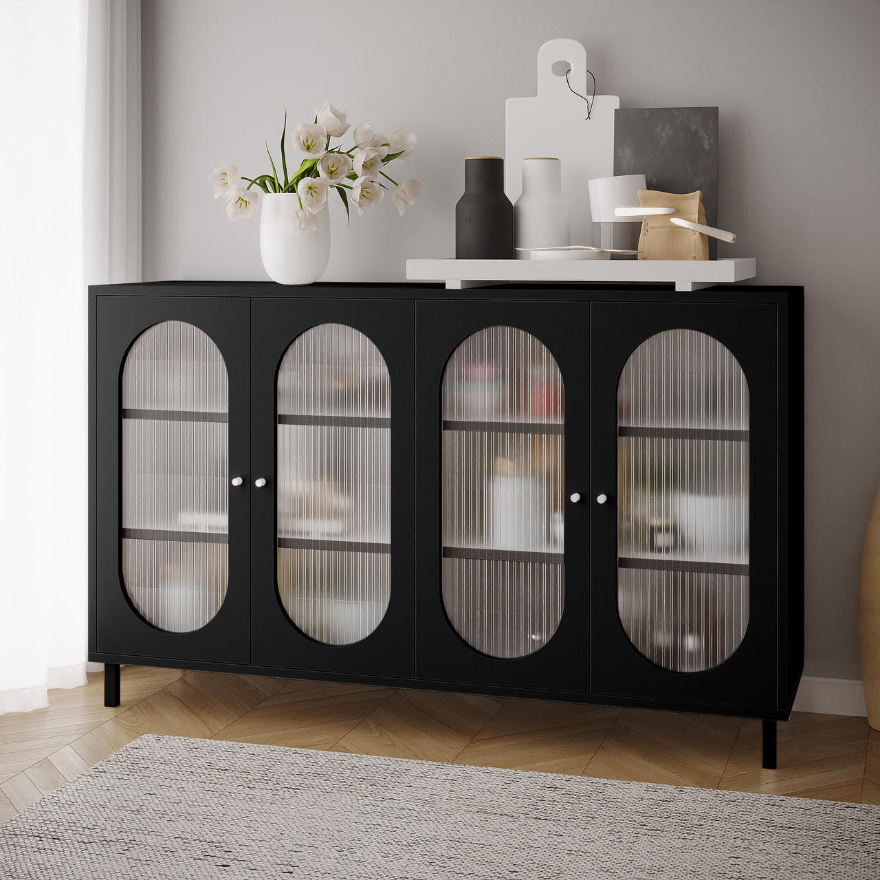 Cambria 150cm Fluted Glass Buffet Cabinet - Black with four ribbed glass doors, topped with a white vase of tulip-like flowers, decorative bottles, and a serving tray on a herringbone floor.