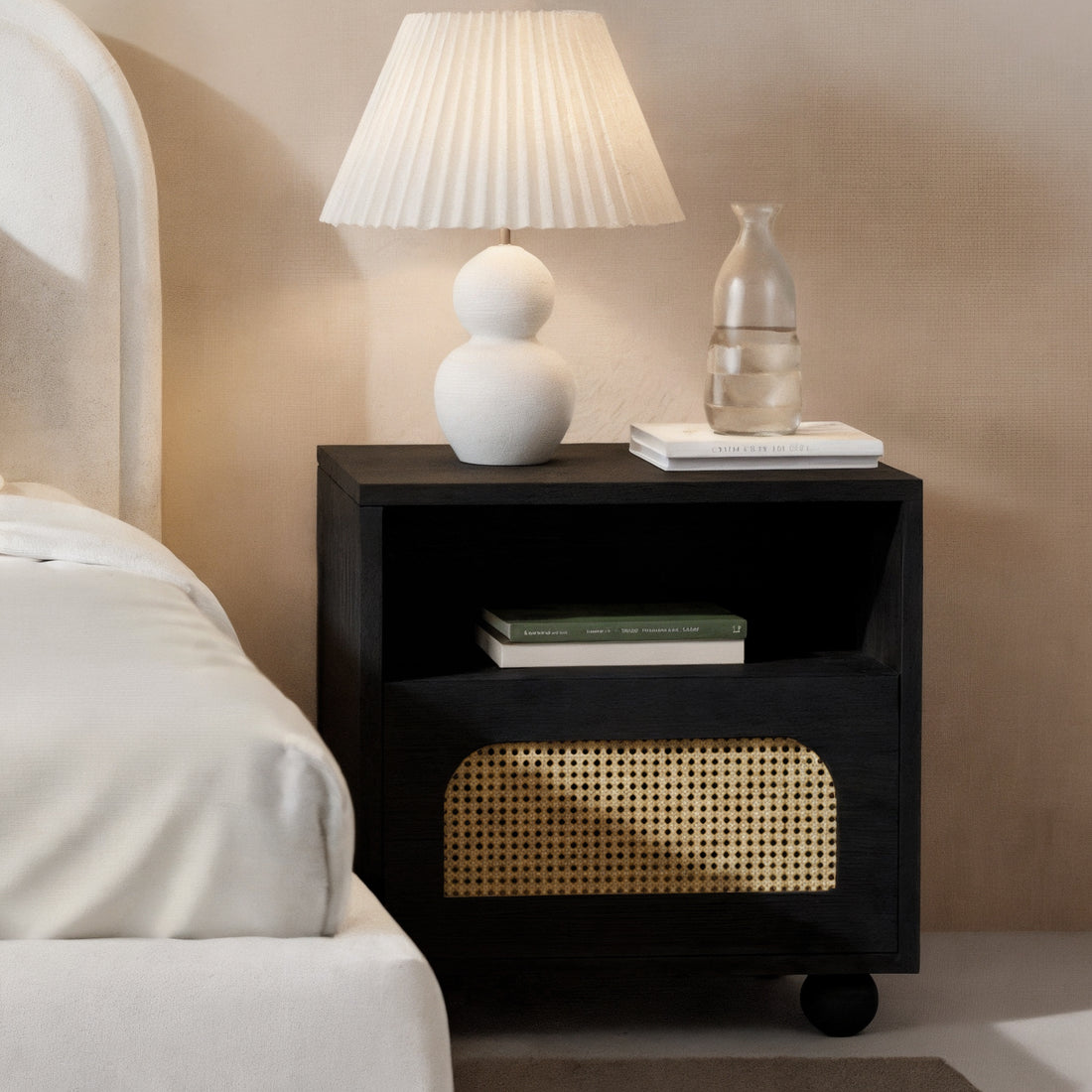 Dialga 45cm Rattan Bedside Table with Storage in Black, featuring a woven cane-front drawer, engineered wood frame, and steel legs, displayed with books, a lamp, and a glass carafe on top.