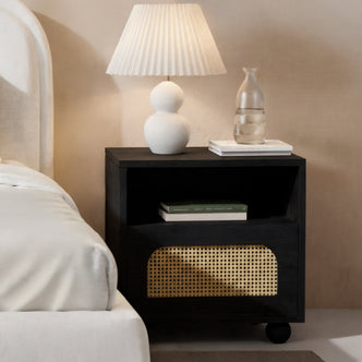 Dialga 45cm Rattan Bedside Table with Storage in Black, featuring a woven cane-front drawer, engineered wood frame, and steel legs, displayed with books, a lamp, and a glass carafe on top.