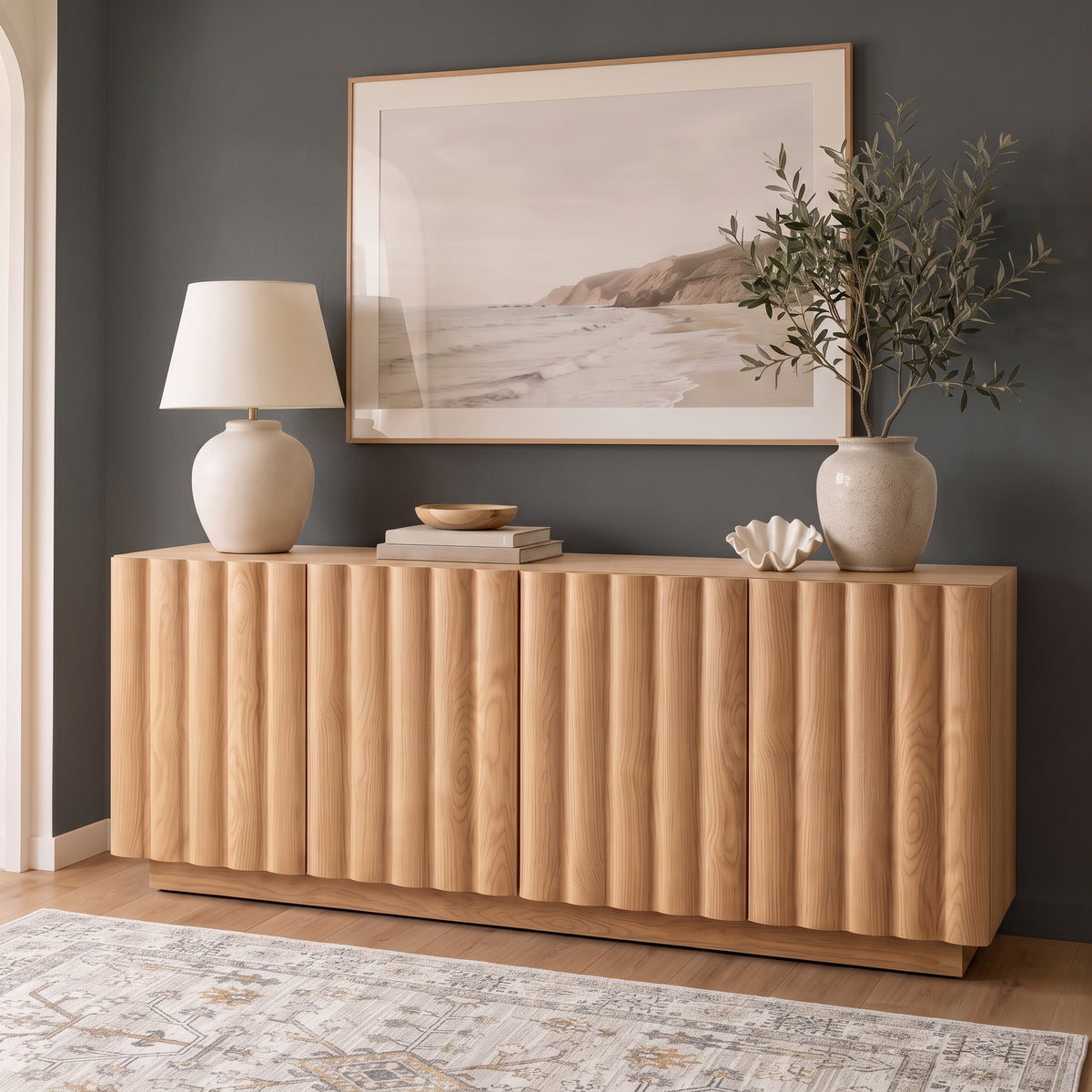 Oak sideboard with vertical fluted panels styled with a ceramic table lamp, potted plant, and framed coastal wall art against a dark grey accent wall.