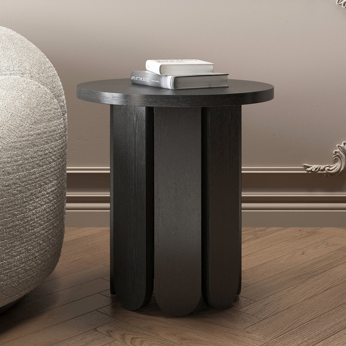 Hugo 45cm Round Side Table with Curve Detail in Black, featuring a scalloped-edge base, is placed on a wood-plank floor beside a boucle-style chair, topped with two stacked books.