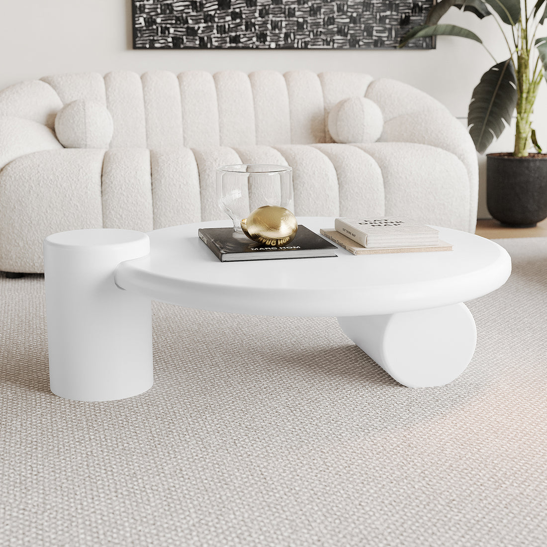Nando 100cm Modern Round Coffee Table in White with two chunky cylindrical legs, featuring stacked books, a double-walled glass vessel, and a golden egg-shaped object on a light woven carpet.