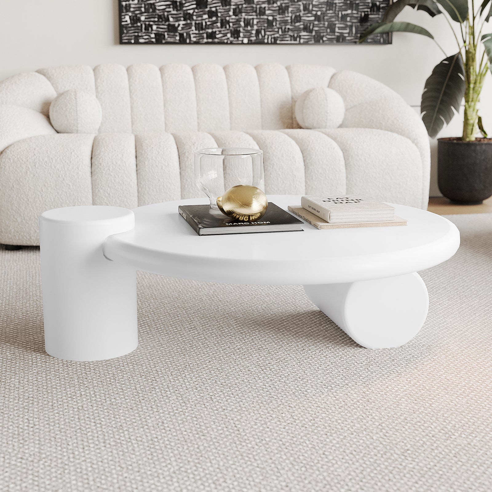 Nando 100cm Modern Round Coffee Table in White with two chunky cylindrical legs, featuring stacked books, a double-walled glass vessel, and a golden egg-shaped object on a light woven carpet.