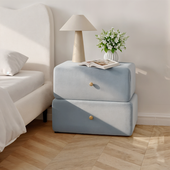 Tyla Velvet Bedside Table with Drawers in Ice Blue beside a bed, featuring a beige lamp, open book, and vase with blossoms, showcasing its velvet finish and compact design.
