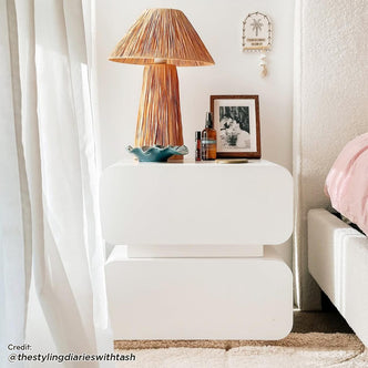 Zimerman 50cm 2-Drawer Bedside Table in White with rounded corners, topped with a woven-straw lamp, teal dish, photo, amber bottles, and coaster; beside a pink-bedded platform and sheer curtain.